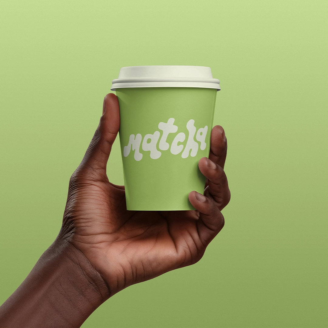 A person holds a paper cup with the word "matcha" printed on it in front of a light green background.